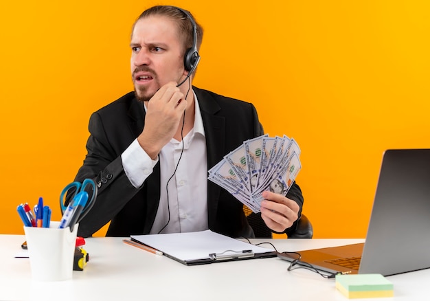 Free photo handsome businessman in suit and headphones with a microphone showing cush looking aside puzzled and displeased sitting at the table in offise over orange background