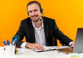 Foto gratuita uomo d'affari bello in vestito e cuffie con un microfono che lavora su un computer portatile che guarda l'obbiettivo sorridente seduto al tavolo in ufficio su sfondo arancione