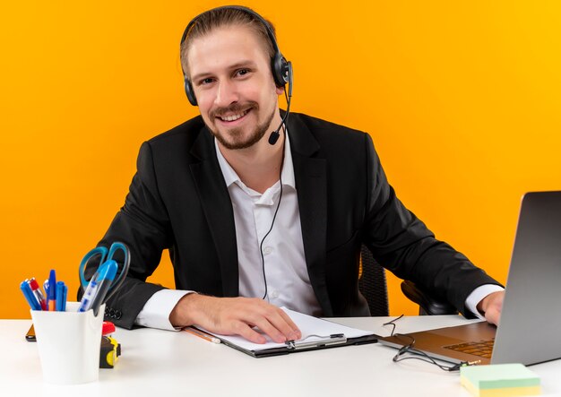 Красивый бизнесмен в костюме и наушниках с микрофоном, работающим на ноутбуке, глядя в камеру, улыбаясь, сидя за столом в офисе на оранжевом фоне