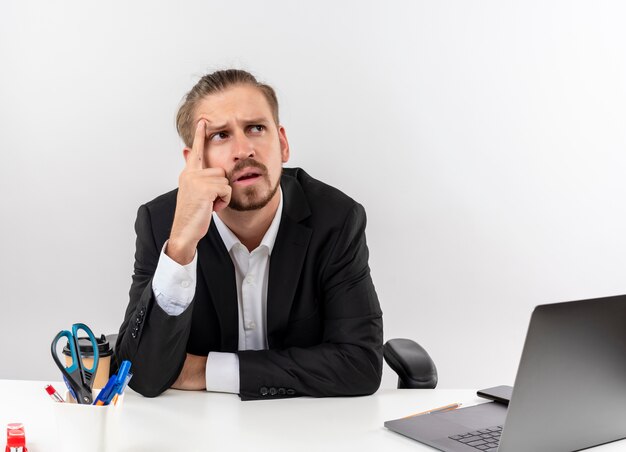 Handsome businessman in suit looking aside puzzled sitting at the table in offise over white background