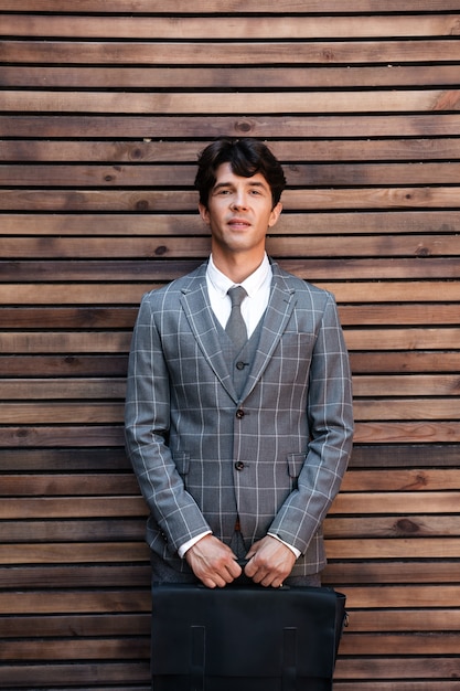 Free photo handsome businessman in suit standing with briefcase against wooden wall