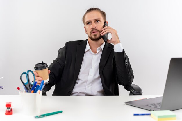 白い背景の上のオフィスでテーブルに座って真面目な顔で脇を見て携帯電話で話しているラップトップに取り組んでいるスーツのハンサムなビジネスマン