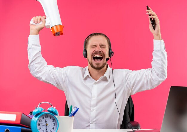 Handsome businessman in white shirt and headphones with a microphone holding megaphone shouting and yelling with aggressive expression sitting at the table in offise over pink background