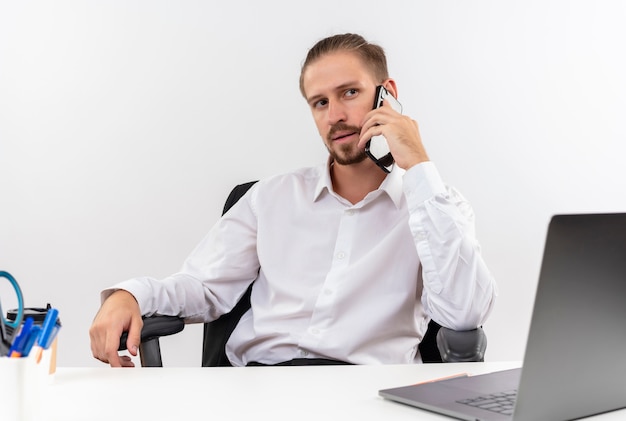 白いシャツとヘッドフォンでハンサムなビジネスマンは、白い背景の上のオフィスでテーブルに座って携帯電話で話している深刻な顔で脇を見てマイクを持っています