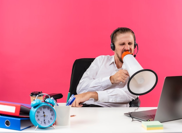 Handsome businessman in white shirt and headphones with a microphone shouting to megaphone with aggressive expression sitting at the table in offise over pink background