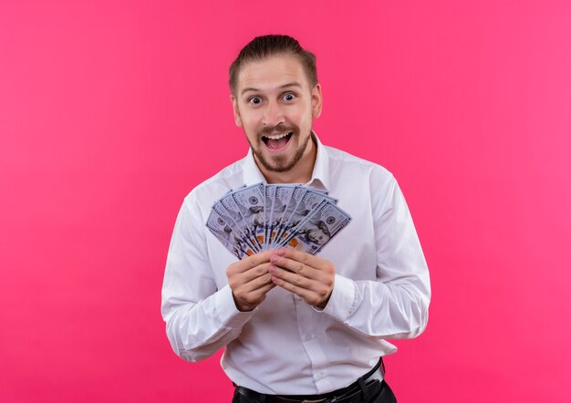 Handsome businessman in white shirt holding cash looking at camera amazed and surprised standing over pink background