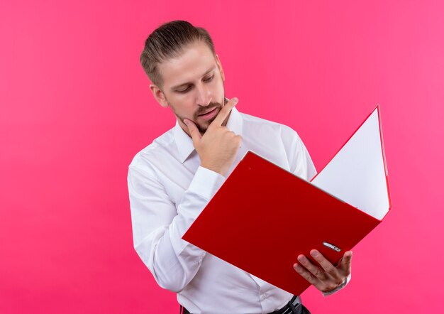 Handsome businessman in white shirt holding folder looking at it with serious face standing over pink background