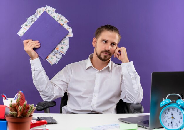 Красивый бизнесмен в белой рубашке, держащий папку с деньгами, глядя в сторону с задумчивым выражением лица, сидит за столом в офисе на фиолетовом фоне