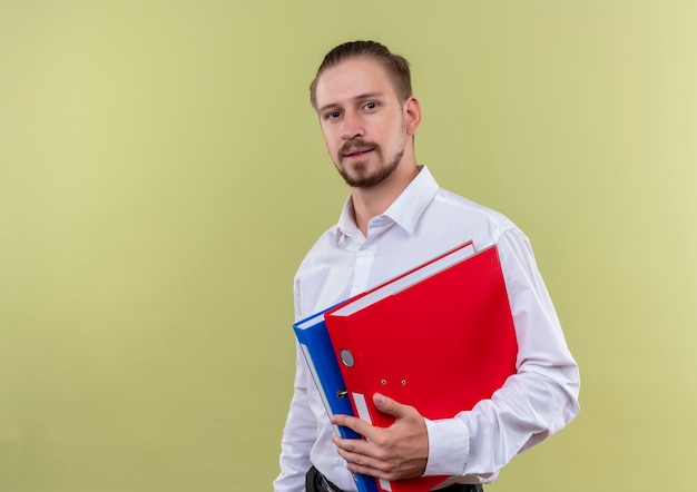 Handsome businessman in white shirt holding folders looking at camera with confident expression standing over olive background