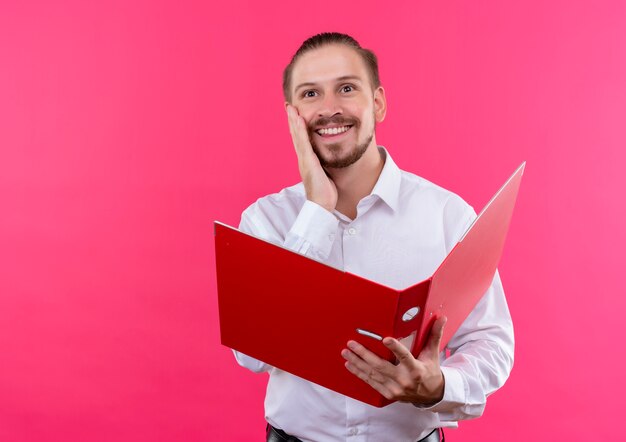 Handsome businessman in white shirt holding open folder looking aside with smile on face feeling positive emotions standing over pink background