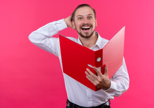 Handsome businessman in white shirt holding open folder looking at camera happy and excited standing over pink background