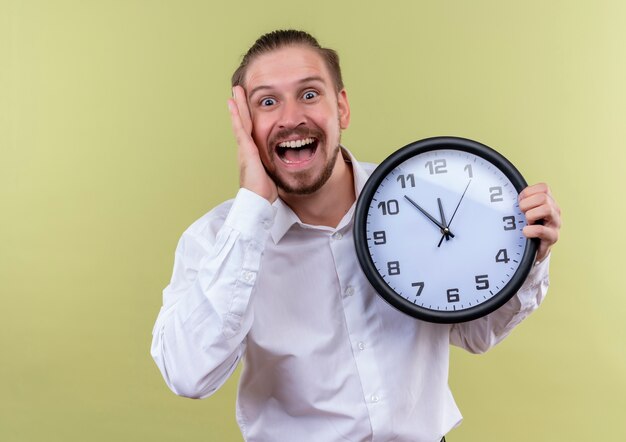 Handsome businessman in white shirt holding wall clock looking at camera surprised and amazed standing over olive background