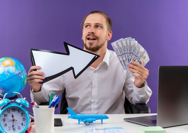 Free photo handsome businessman in white shirt holding white arrow showing cash looking aside with smile on face sitting at the table in offise over purple background