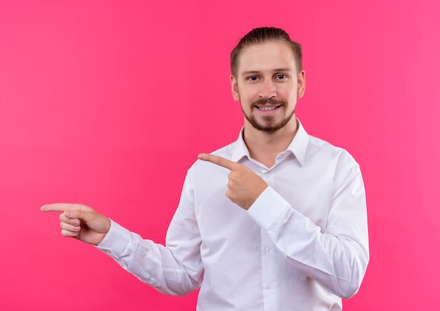 Foto gratuita uomo d'affari bello in camicia bianca che guarda l'obbiettivo sorridente che punta con il dito indice a lato in piedi su sfondo rosa