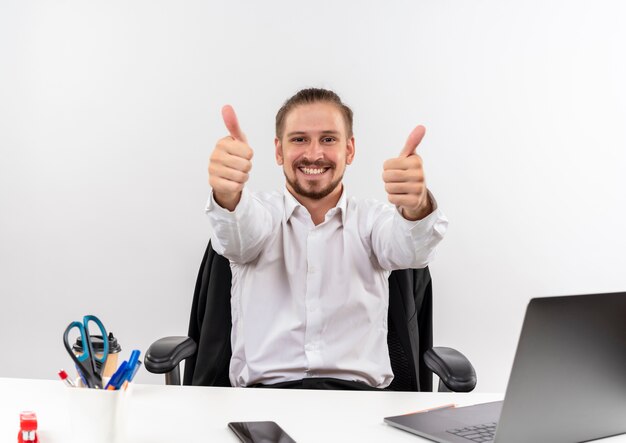 Handsome businessman in white shirt looking at camera smiling showing thumbs up sitting at the table in offise over white background