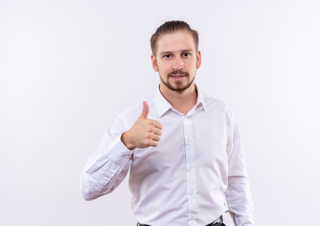 Handsome businessman in white shirt looking at camera smiling showing thumbs up standing over white background