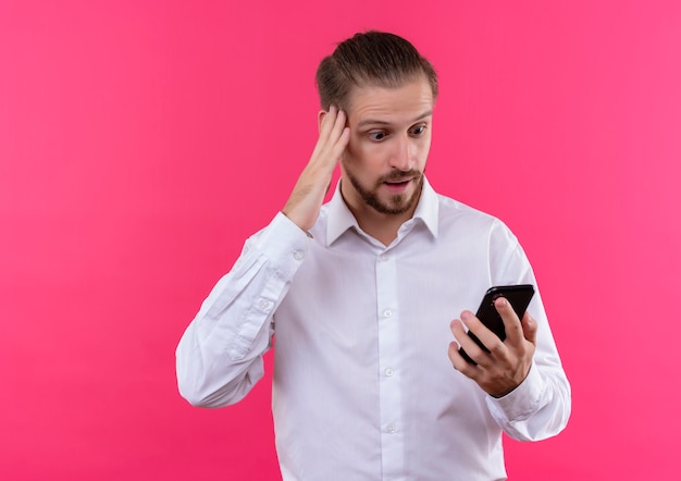 Handsome businessman in white shirt looking at screen of his smartphone surprised and amazed standing over pink background