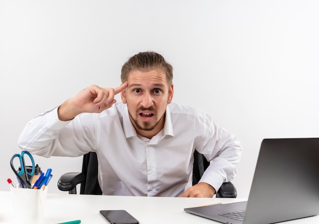 Handsome businessman in white shirt pointing finger his temple looking at camera displeased sitting at the table in offise over white background