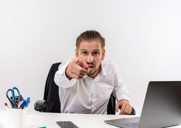 Handsome businessman in white shirt pointing with index finger to camera displeased sitting at the table in offise over white background