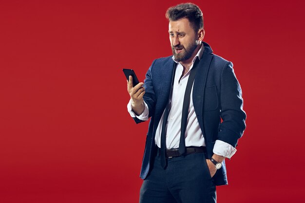 Handsome businessman with mobile phone. Happy business man standing isolated on red studio background. Beautiful male half-length portrait. Human emotions, facial expression concept.