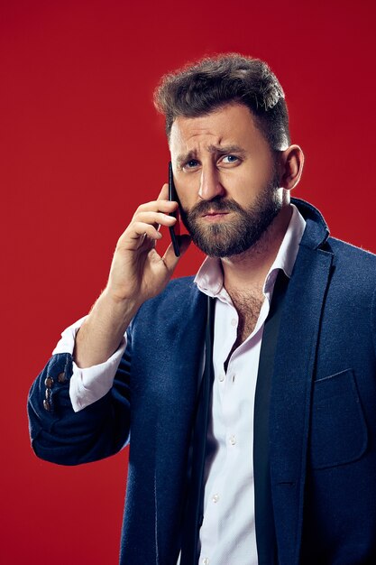 Handsome businessman with mobile phone. Serious business man standing isolated on red  wall. Beautiful male half-length portrait. Human emotions, facial expression concept.