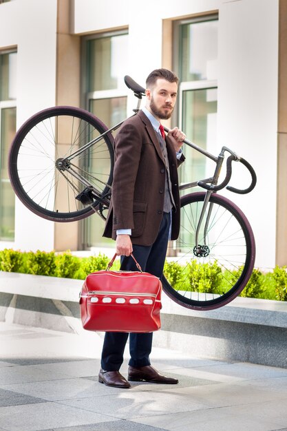 街の通りで彼の自転車を運ぶ赤いバッグでハンサムな実業家。若い男性の現代的なライフスタイルの概念