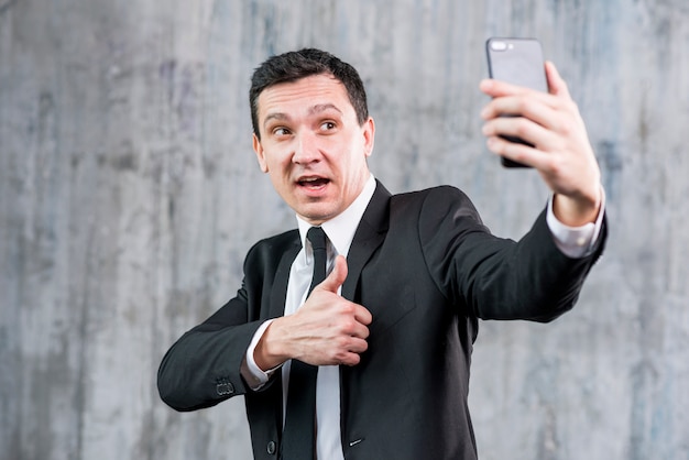 Uomo d'affari bello con il pollice in su prendendo selfie