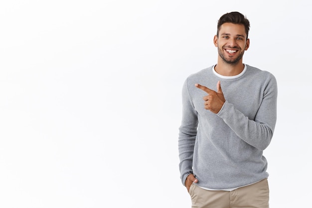 Handsome carefree bearded caucasian man in grey sweater pointing left discuss product smiling as talking to you with friendly expression showing around introduce promo banner white background