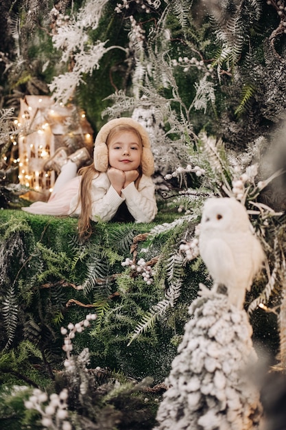 긴 공정한 머리를 가진 잘 생긴 백인 아이는 그녀와 올빼미 주위에 많은 장식 나무와 함께 크리스마스 분위기에 놓여 있습니다