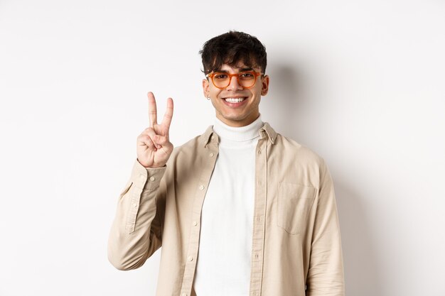 Handsome caucasian guy in glasses showing two fingers and smiling, standing on white background