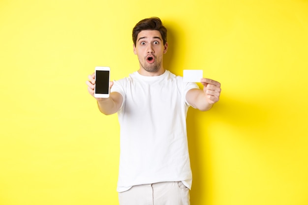 Handsome caucasian guy showing smartphone screen and credit card, concept of mobile banking and online shopping, yellow background.