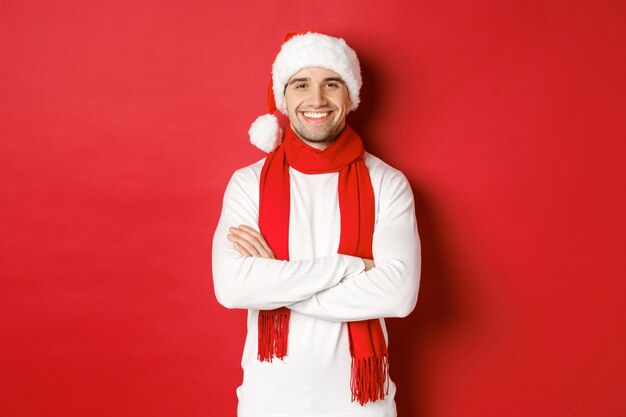 Handsome caucasian guy with bristle wearing santa hat scarf and white sweater cross arms on chest an...