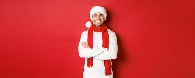 Handsome caucasian guy with bristle wearing santa hat scarf and white sweater cross arms on chest an...