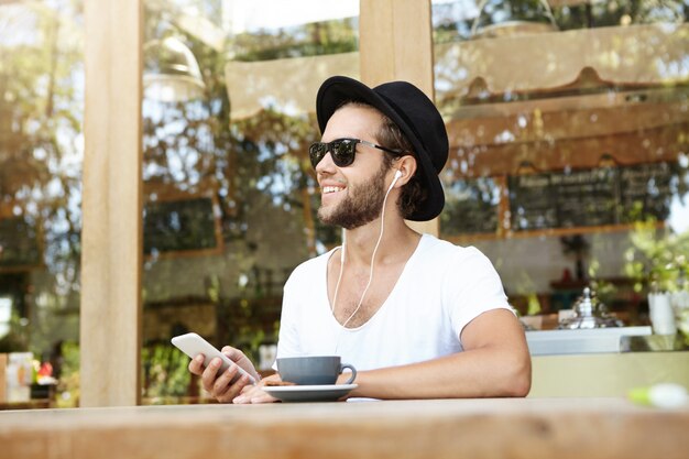 Handsome Caucasian hipster wearing trendy hat and sunglasses enjoying free wifi at coffee shop