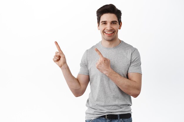 Handsome caucasian man smiling, pointing upper left corner, showing promo text aside on copyspace, standing in casual clothes against white background