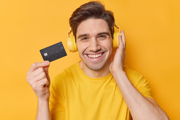 Handsome cheerful adult man holds credit card spends money for online shopping listens music via headphones dressed in casual t shirt isolated over yellow background People and finances concept