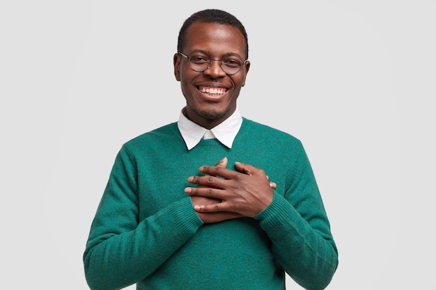 Handsome cheerful black man keeps both hands on chest, feels touched or thankful, smiles broadly, wears elegant green sweater