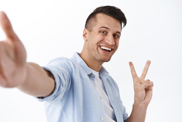 Handsome cheerful caucasian man taking selfie on smartphone hold camera and show peace sign smiling carefree make photo with mobile phone during his travel abroad staying in touch