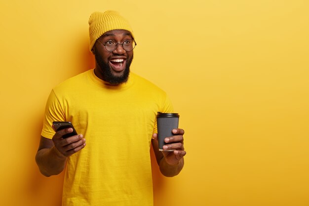 Handsome cheerful dark skinned guy with thick bristle, has fun indoor, holds cellular, waits for call, enjoys drinking hot beverage, isolated over yellow wall. People, technology concept