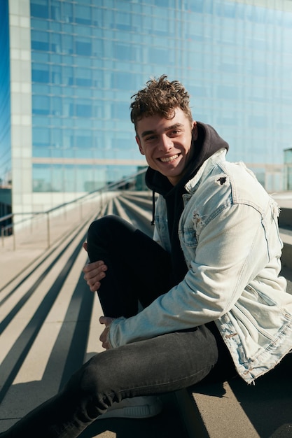 Handsome cheerful guy in denim jacket and hoodie happily looking in camera resting on city stairs