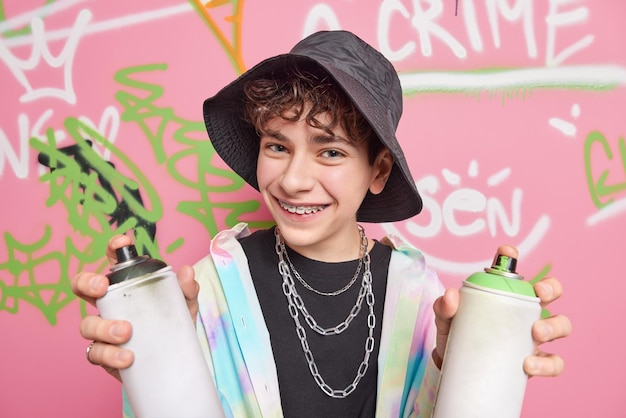 Handsome cheerful hipster boy poses in city against graffiti background wears black panama shirt silver chains braces on teeth holds two cans of paint enjoys favorite hobby Street art concept