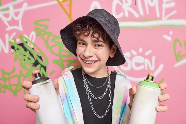 Handsome cheerful hipster boy poses in city against graffiti background wears black panama shirt silver chains braces on teeth holds two cans of paint enjoys favorite hobby Street art concept