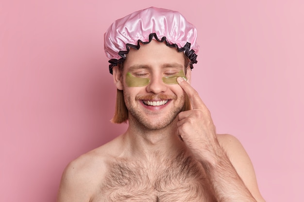 Handsome cheerful man closes eyes points at hydrogel patches undergoes skin care treatment