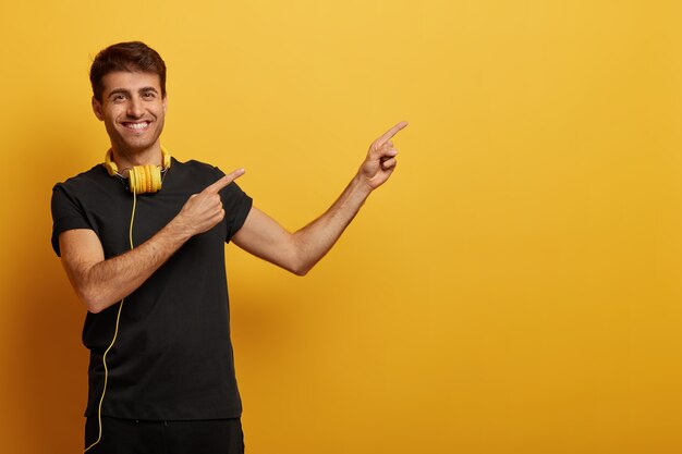 Handsome cheerful man points at copy space, dressed in black clothes, wears headset, smiles toothily, demonstrates advertisement, isolated over yellow background