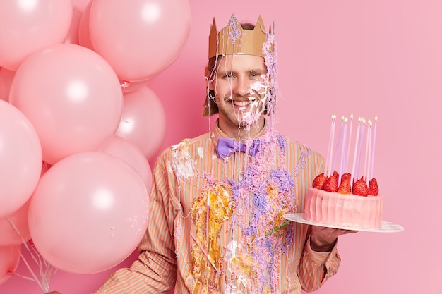 Красивый веселый мужчина стоит грязно со сливками и держит вкусный клубничный торт празднует день рождения с праздничными атрибутами, изолированными над розовой стеной