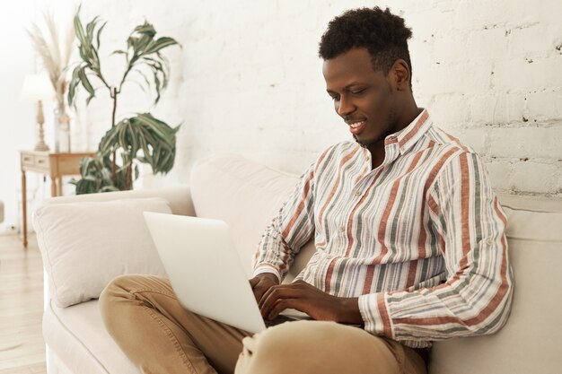 ラップトップコンピューターでソファに座って自宅で仕事を楽しんでいるハンサムな陽気な若い暗い肌の男性のsmmマネージャー