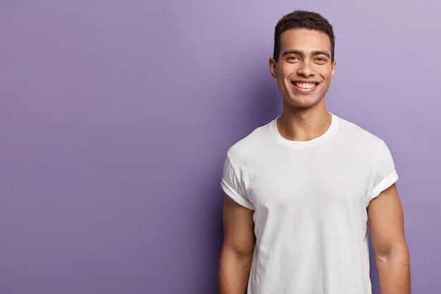 ハンサムで陽気な若いスポーツマンは、スポーティな体、筋肉質の腕、白いモックアップTシャツを着て、短い黒髪、歯を見せる魅力的な笑顔、紫色の壁の上に立って、空白のコピースペースを脇に置いています