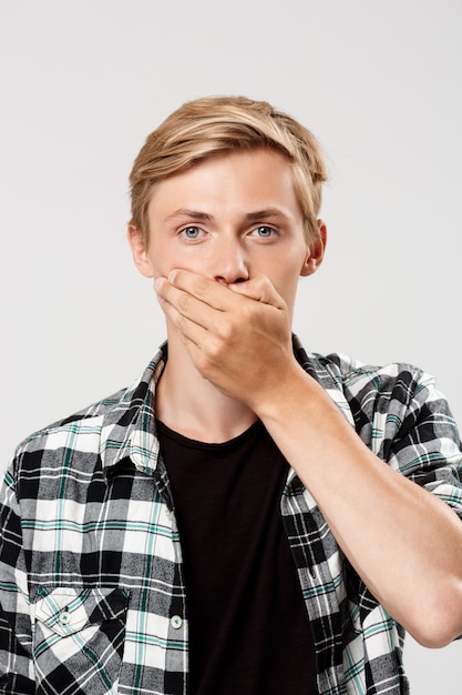  handsome confident blond young man wearing casual plaid shirt covering mouth with hand, on grey wall