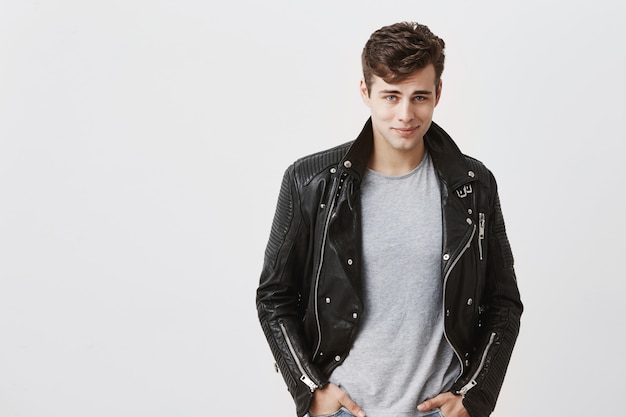 Handsome confident good-looking male model dressed in trendy black leather jacket over gray t-shirt, looking  with his blue appealing eyes, posing .
