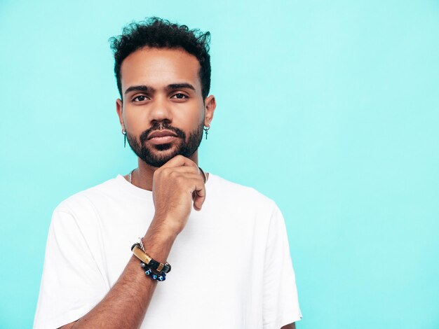 Free photo handsome confident hipster model sexy unshaven man dressed in white summer tshirt and jeans clothes fashion male posing near blue wall in studiolooking at camera and touching his chin